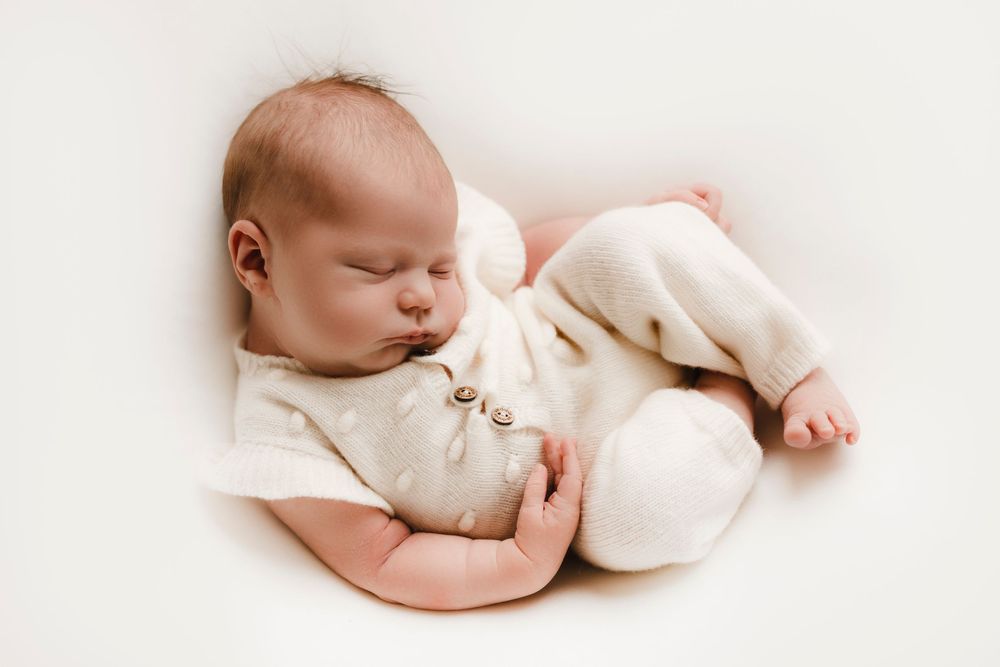 Peaceful newborn baby sleeping curled up in cream outfit on white background.