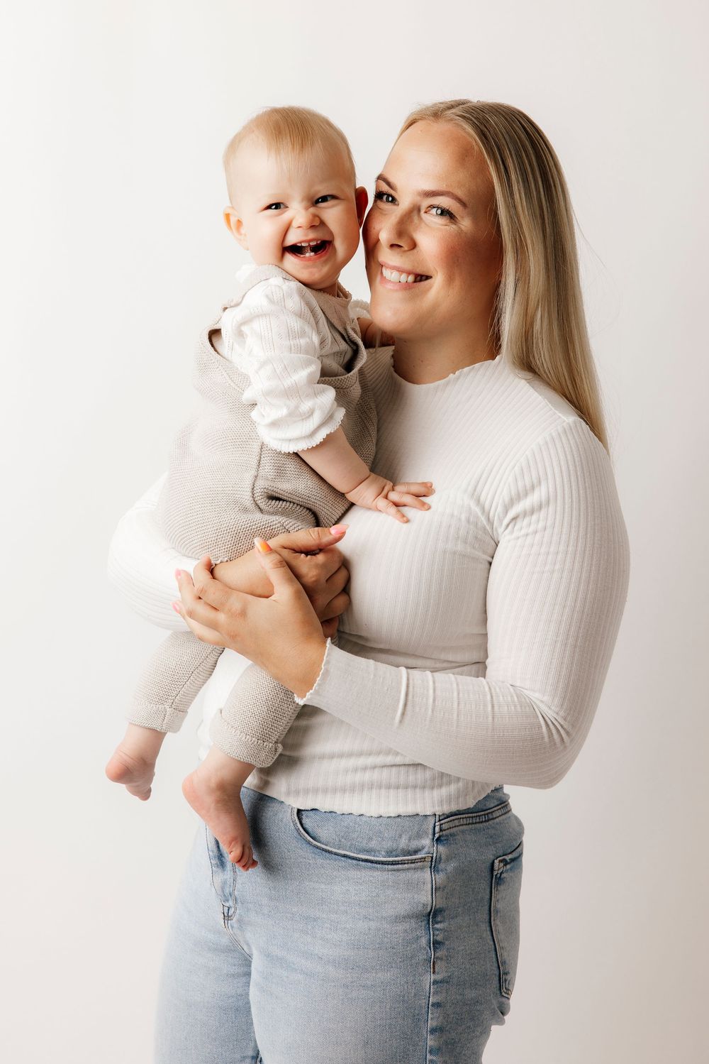 Smiling blonde woman holding a laughing baby in gray outfit against a white background.