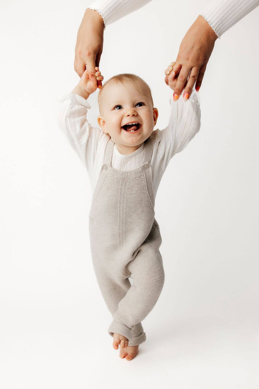 Smiling baby in gray overalls and white shirt learning to walk with adult hands supporting raised arms on white background.