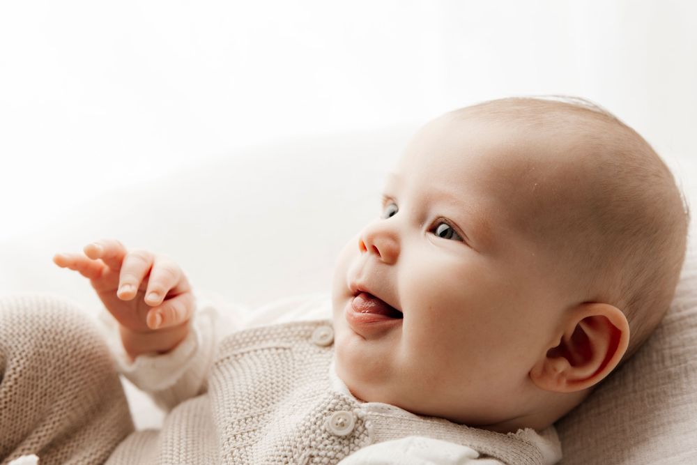 Smiling baby lying on back in a knit onesie, looking up with curiosity and reaching one hand upward.