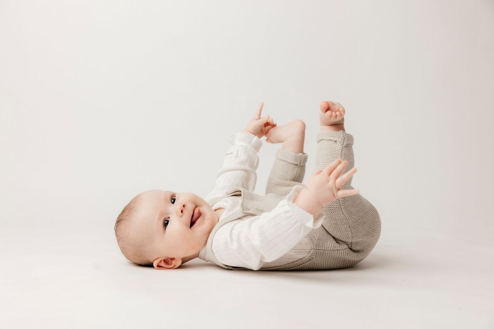 A happy baby lying on its back on a white background, wearing a gray outfit, smiling and lifting its legs.