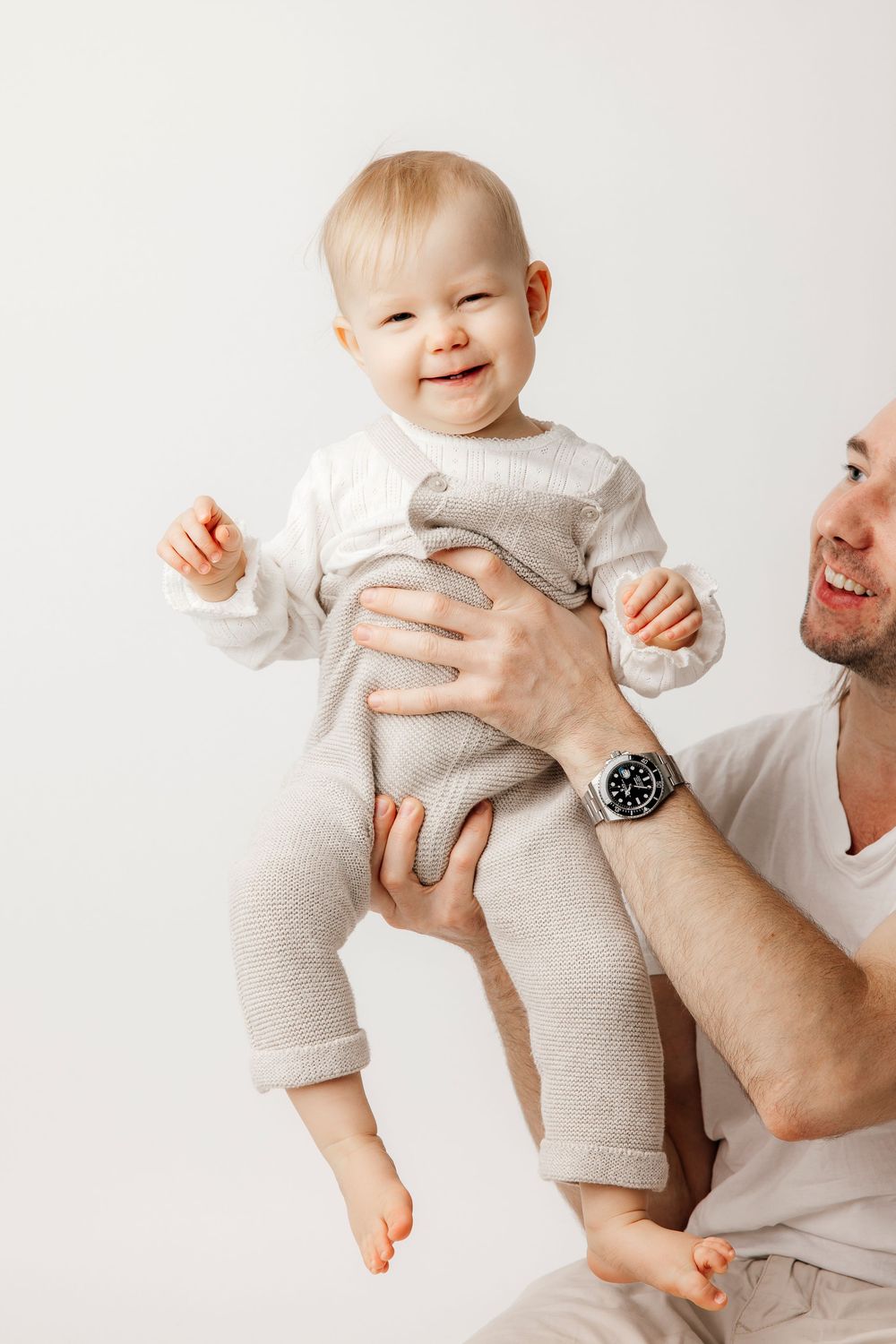 Happy baby in beige onesie being held up by smiling father against white background.