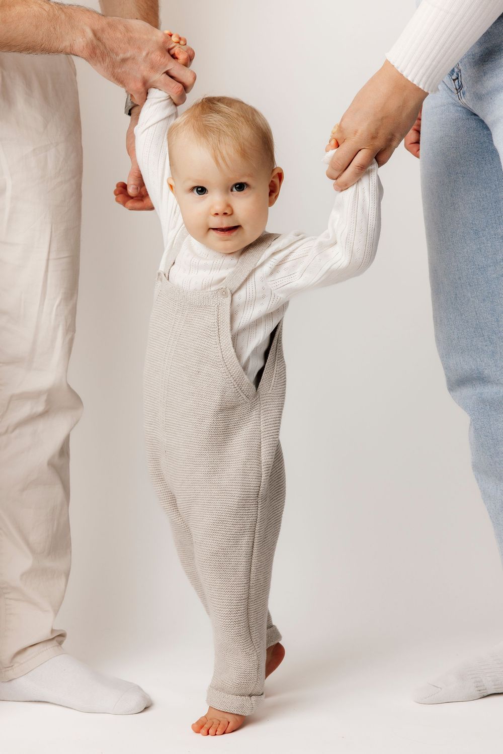 A smiling baby in a gray romper takes first steps while being supported by two adults holding their hands.