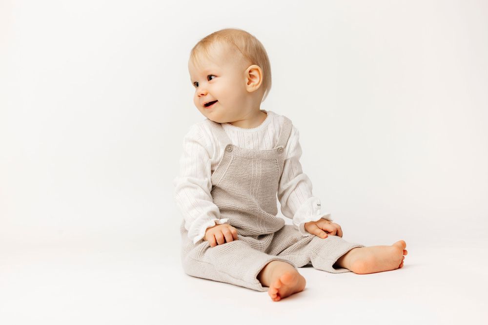 Adorable baby sitting on white background wearing linen overalls and white long-sleeve shirt, looking sideways.