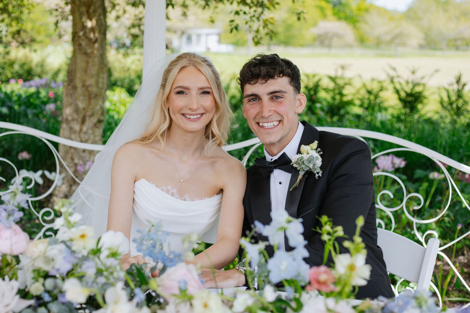 Smiling bride and groom seated at floral-decorated wedding table outdoors, surrounded by pink and white blooms.