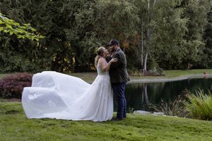 Bride and groom share a romantic moment by a pond, with her white gown flowing in the wind, surrounded by lush greenery.