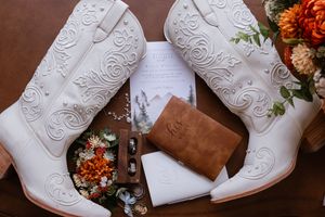 White cowboy boots with floral embroidery displayed with wedding stationery, rings, and bouquet on a wooden surface.