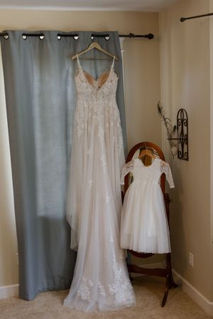 Elegant lace wedding gown hanging beside a matching flower girl dress on a wooden chair in a bridal suite.