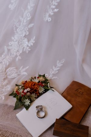 Wedding rings rest on vow books beside autumn floral bouquet and lace bridal gown backdrop.