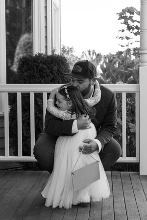 Black and white photo of a man in a cap hugging a young flower girl in a white dress on a wooden porch.