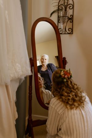 Bride with floral hair piece looks into ornate wooden mirror as older woman helps her prepare for wedding day.