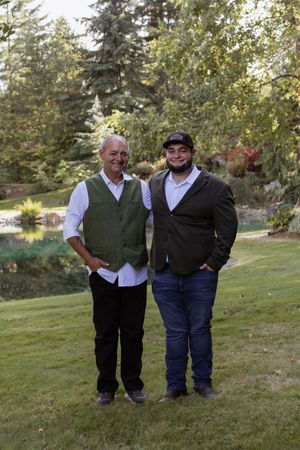 Two men in green blazers smile outdoors on a lawn surrounded by trees and a pond.