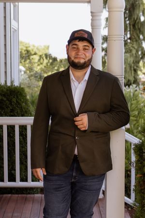 Man in dark blazer, white shirt, and baseball cap smiling on a porch with white columns and greenery.