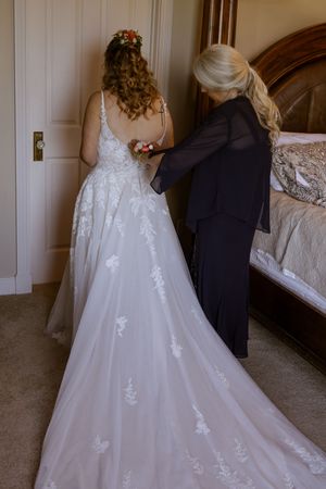 A bride in a white lace wedding gown gets her dress fastened by a woman in black in an elegant bedroom.