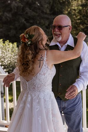 Bride in lace wedding dress with cross tattoo dances with bald older man in green vest at outdoor wedding.