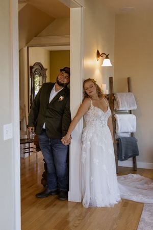 Bride in white lace gown and groom in green jacket hold hands in a doorway of a warmly lit home.