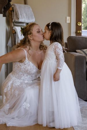 Bride in lace wedding gown shares a sweet kiss with flower girl in white tulle dress during wedding preparations.