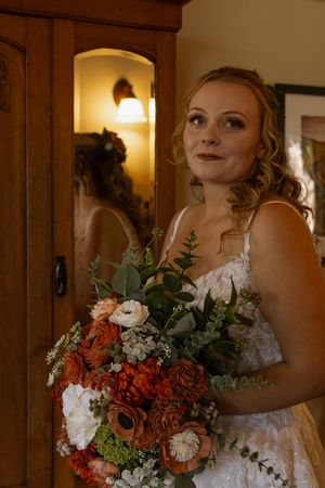 Bride holding a lush bouquet of rust, white, and green flowers, standing before a mirror in warm indoor lighting.