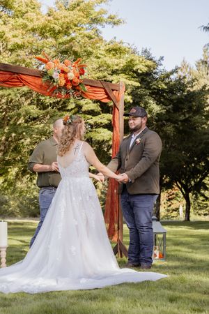 Bride and groom hold hands at outdoor wedding ceremony beneath rustic wooden arch with orange floral arrangements.