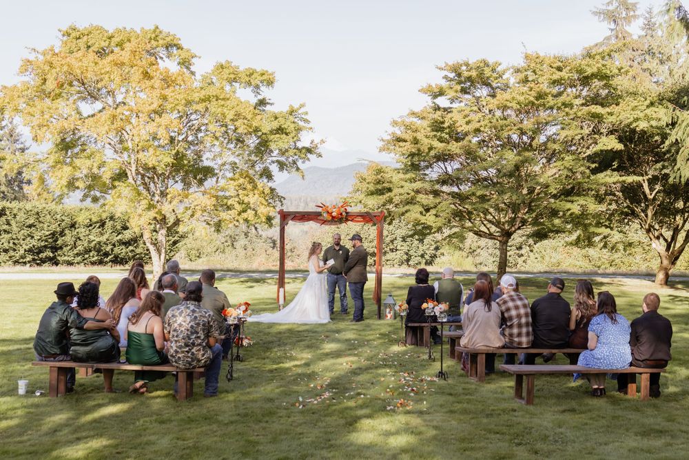 Outdoor wedding ceremony on green lawn with guests seated on wooden benches facing couple at rustic arch amid autumn trees.