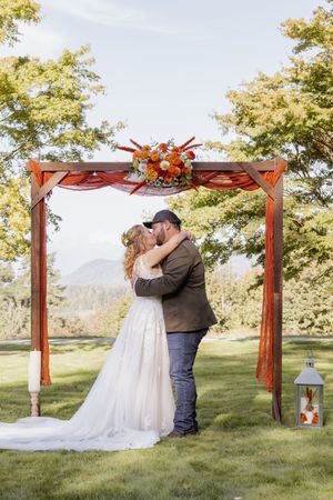Bride and groom share first kiss under rustic wooden arch with fall floral decor outdoors.