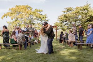 Couple shares first kiss at outdoor wedding ceremony with guests standing beside rustic wooden benches under green trees.