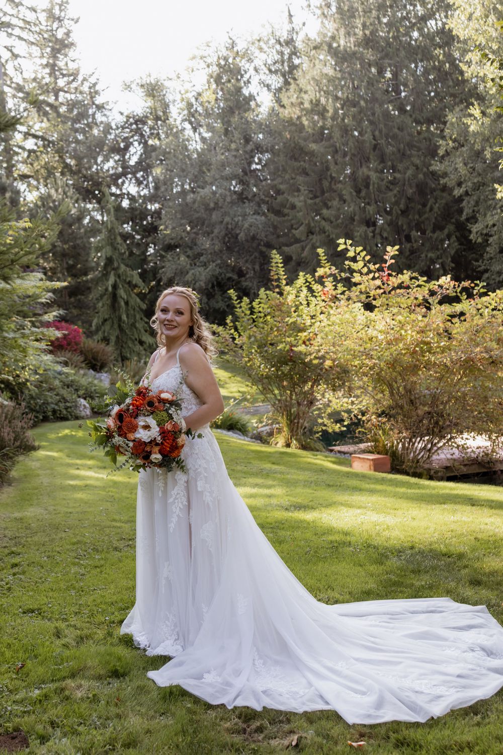 Radiant bride in elegant white lace gown with long train holds red bouquet while posing in lush garden.