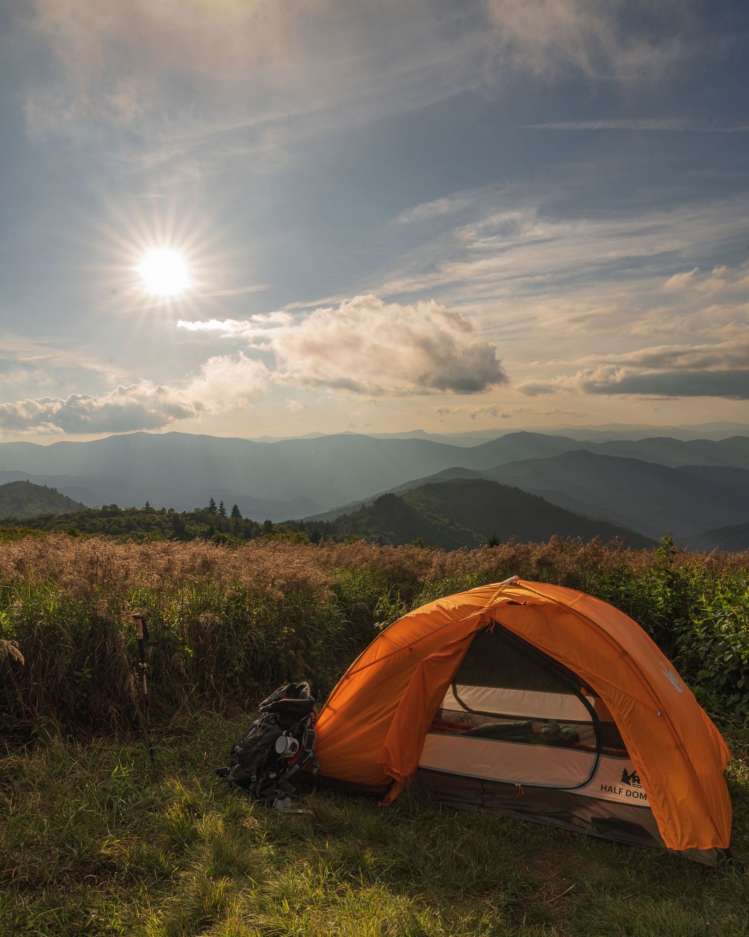 Orange camping tent set up on a mountain meadow with panoramic views of the Blue Ridge Mountains of WNC under a bright sunny sky.