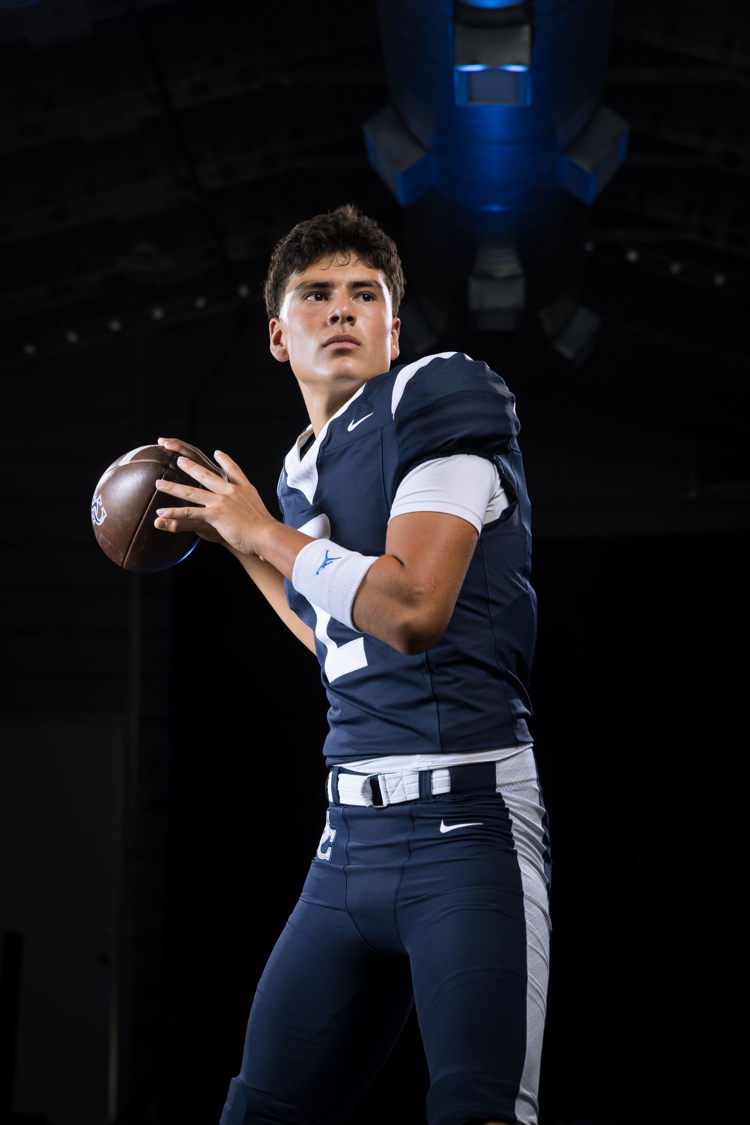 A young football player in navy blue Nike uniform holds a football in throwing position against a dark dramatic background.