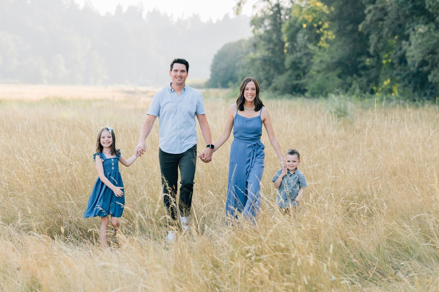 Family of four walking through golden field, parents holding children's hands, surrounded by tall grass and trees.
