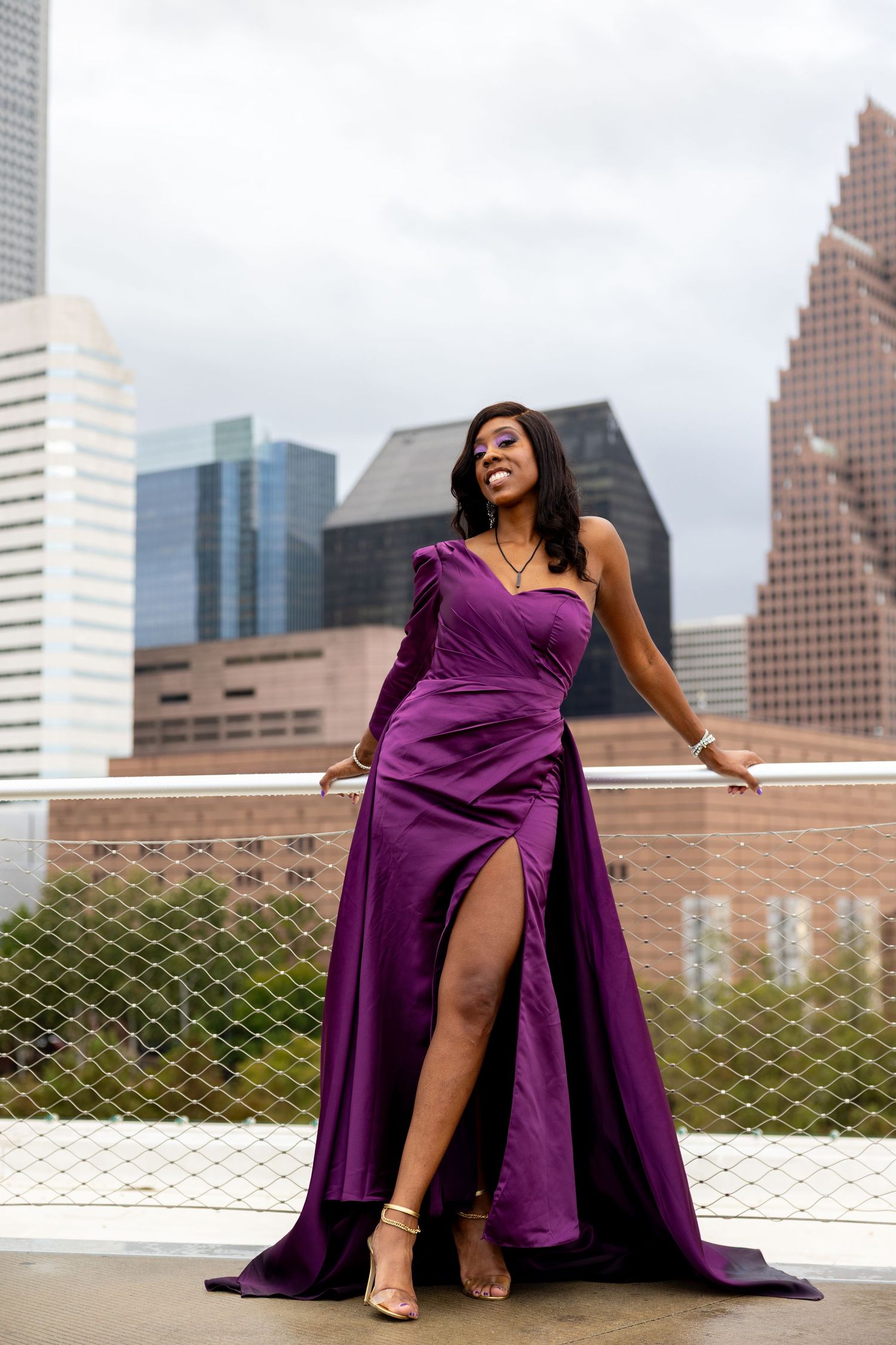 Woman in purple one-shoulder satin gown with high slit poses on urban rooftop with city skyline backdrop.