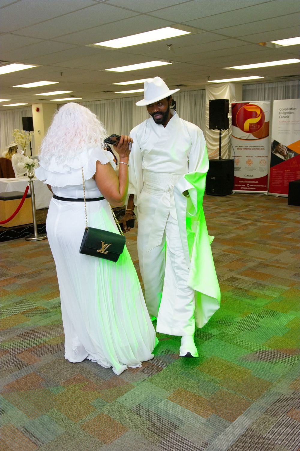 Two elegantly dressed individuals in white outfits at a formal event, man wearing white hat and woman carrying Louis Vuitton bag.