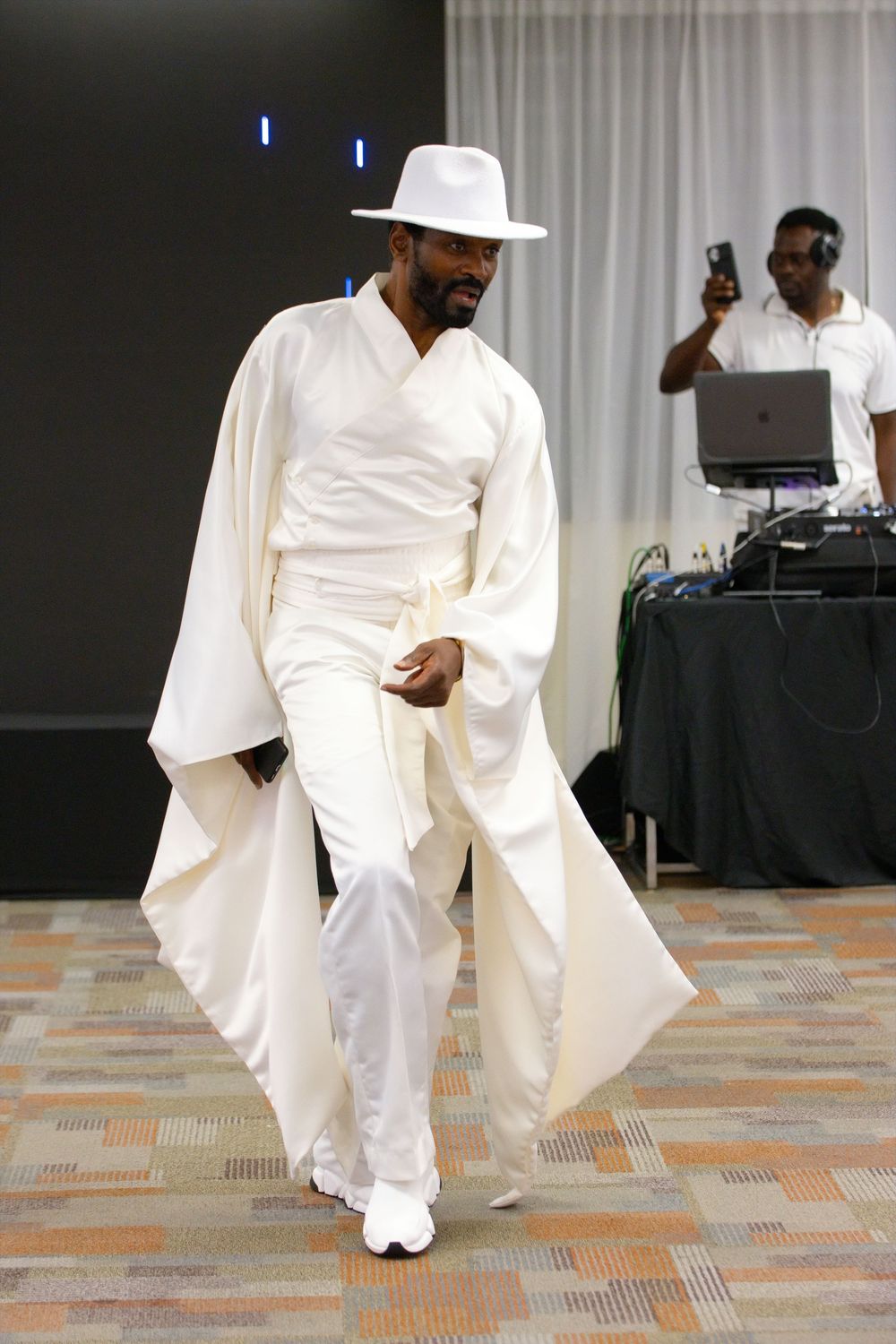 Man in all-white ensemble with wide-brimmed hat and flowing cape walks runway at fashion show.