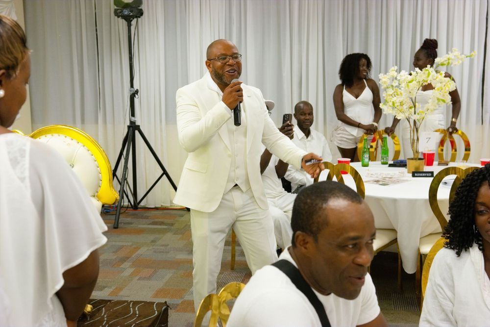 Man in white suit speaks into microphone at elegant white and gold themed party event.