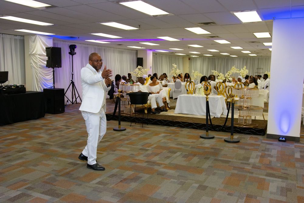 Man in white suit speaks at elegant banquet hall event with guests seated at decorated tables.