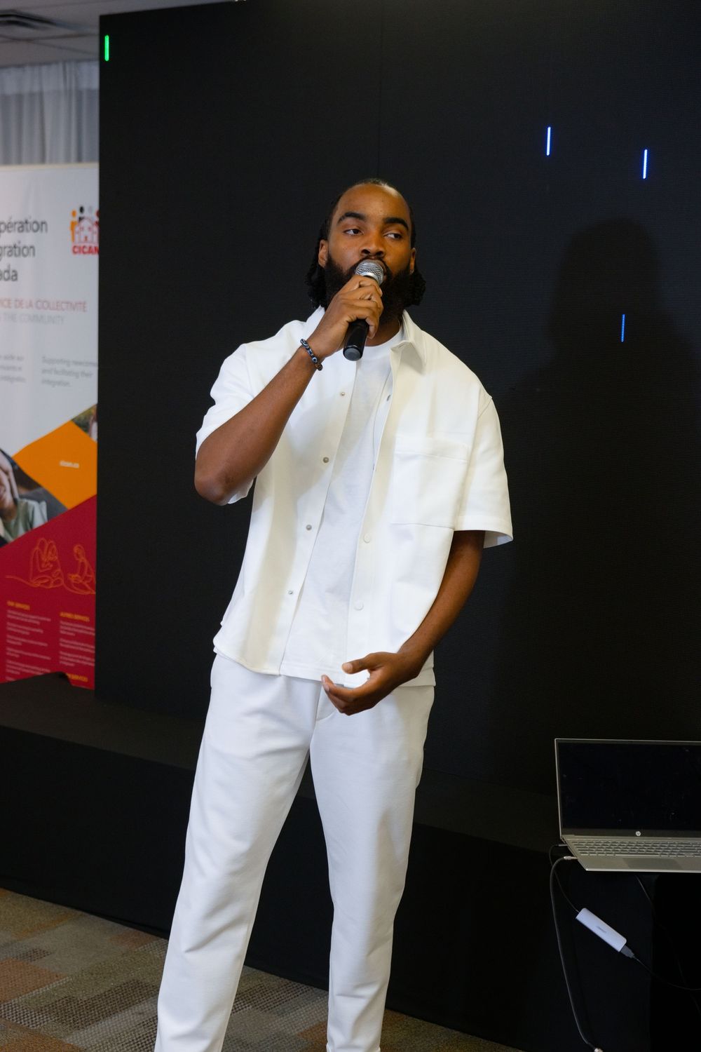 Man in white outfit speaking into microphone at indoor event with promotional banners in background.