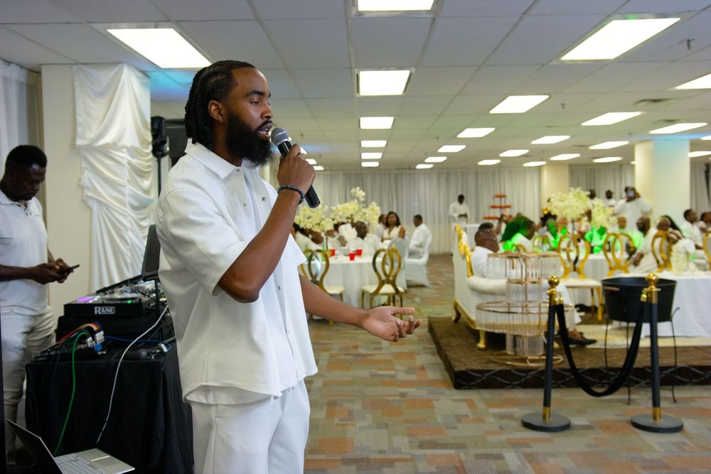 Man in white outfit speaks into microphone at elegant event with guests seated in decorated banquet hall.