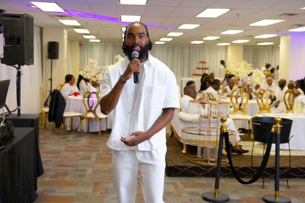 Man in white outfit speaks into microphone at elegant banquet hall event with decorated tables in background.