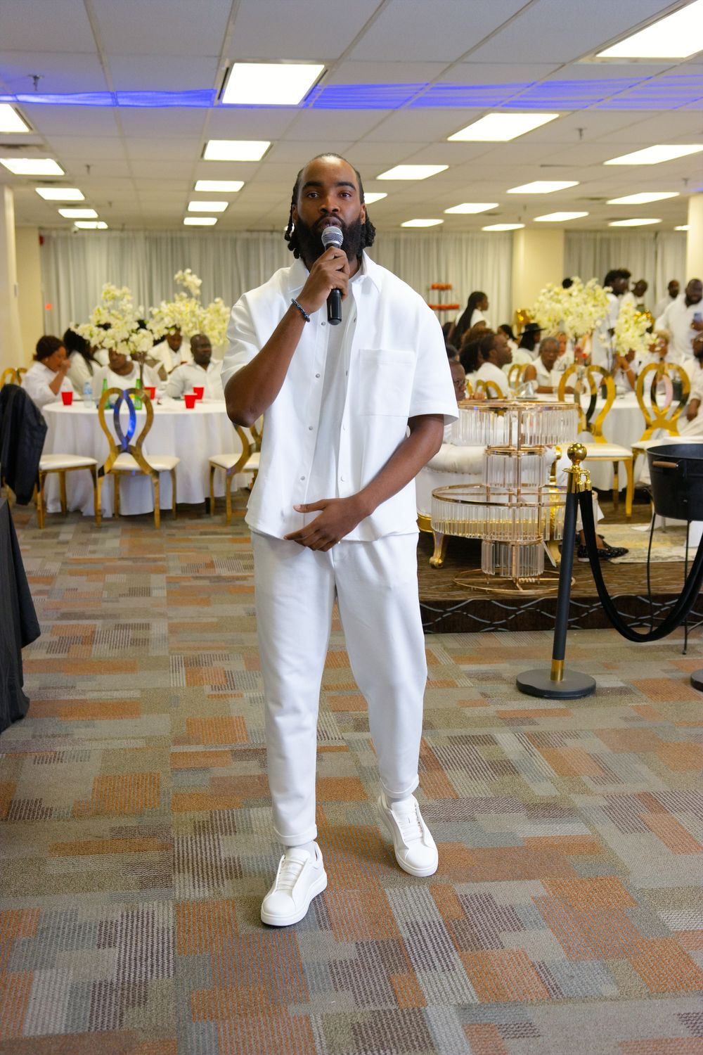 Man in all-white outfit speaks into microphone at elegant banquet hall event with gold decor.
