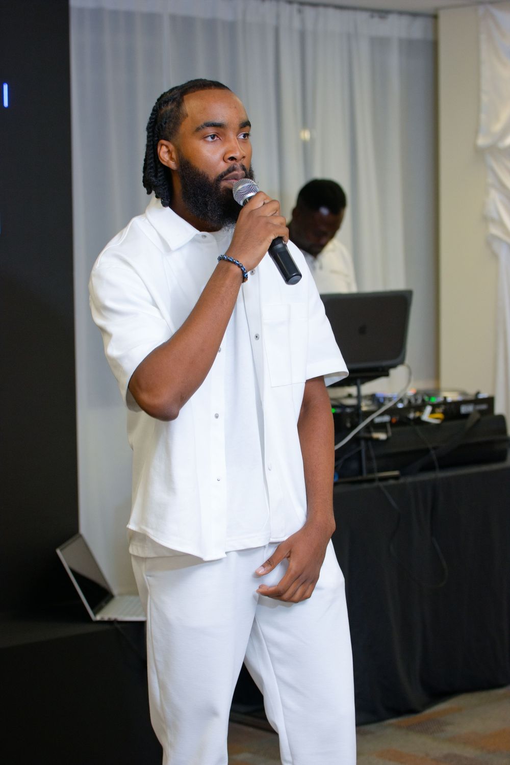 Man in white outfit singing into microphone at indoor event with DJ equipment visible in background.