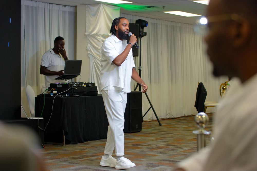 Man in white outfit speaking into microphone at indoor event with DJ setup in background.