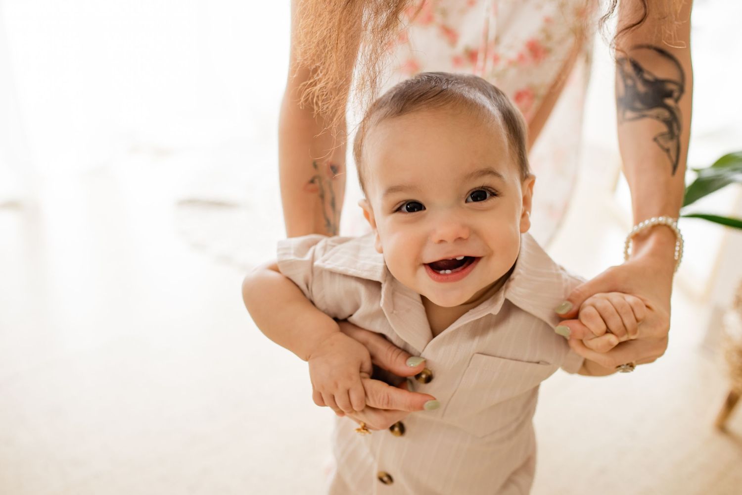 Smiling baby in beige button-up shirt being supported by tattooed adult hands in bright airy room.