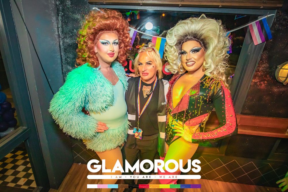 Three people pose together at a Glamorous drag event, two in colorful drag costumes with big hair flanking a person in casual attire.