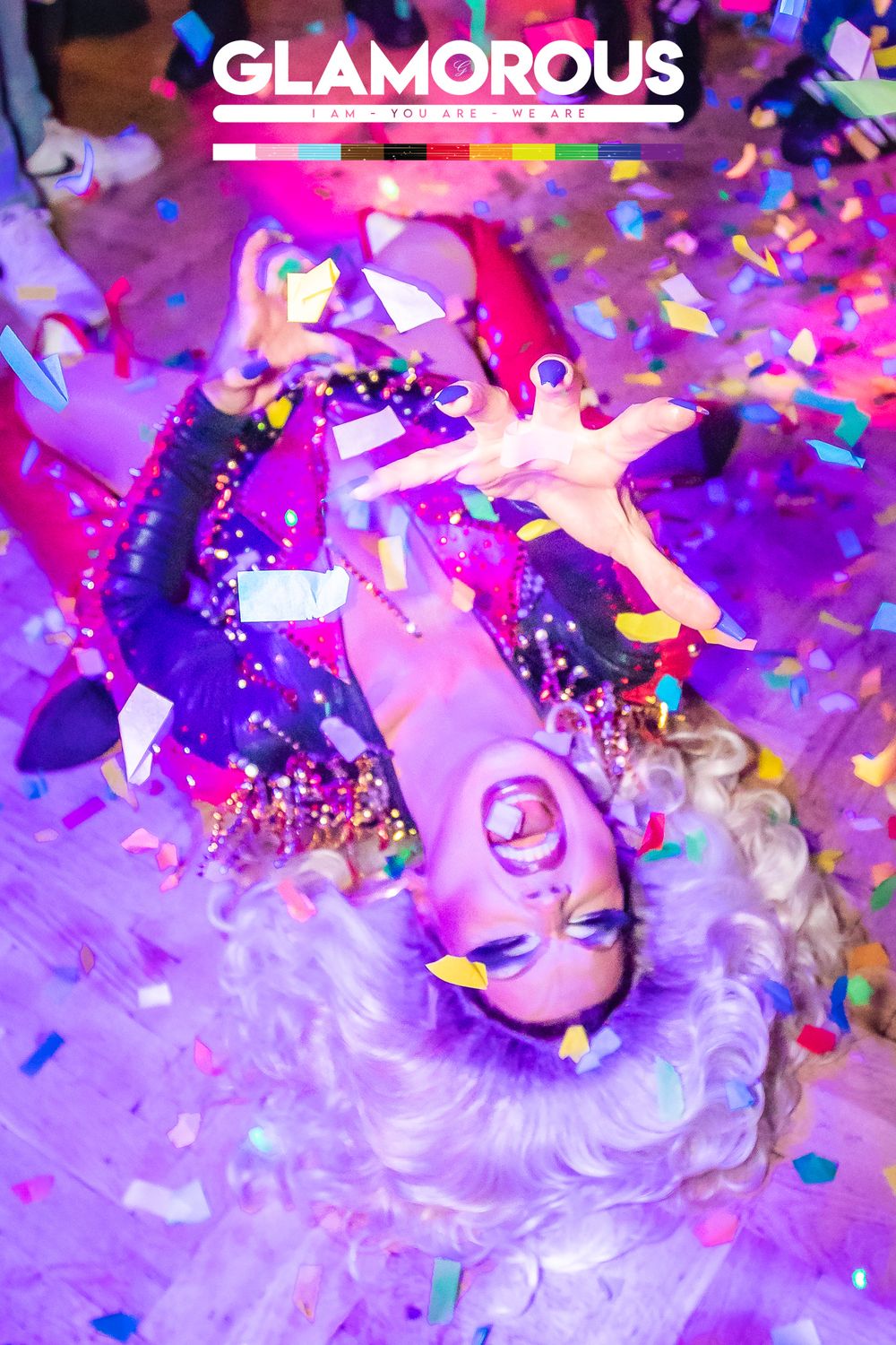 Drag performer lying on floor covered in colorful confetti under purple lights at Glamorous club event.