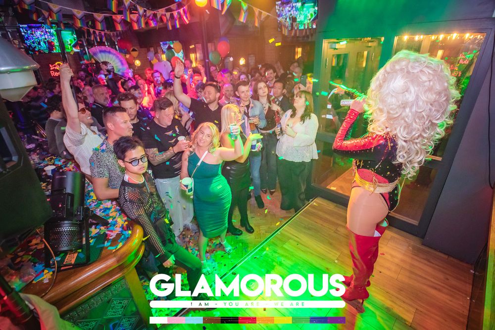 Drag queen performer entertaining a lively crowd at Glamorous nightclub event with colorful lights.