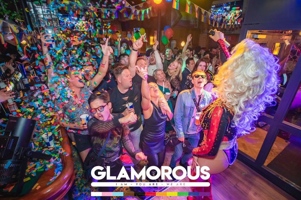 Glamorous drag queen performing at a lively LGBTQ+ nightclub party with confetti and dancing crowd.