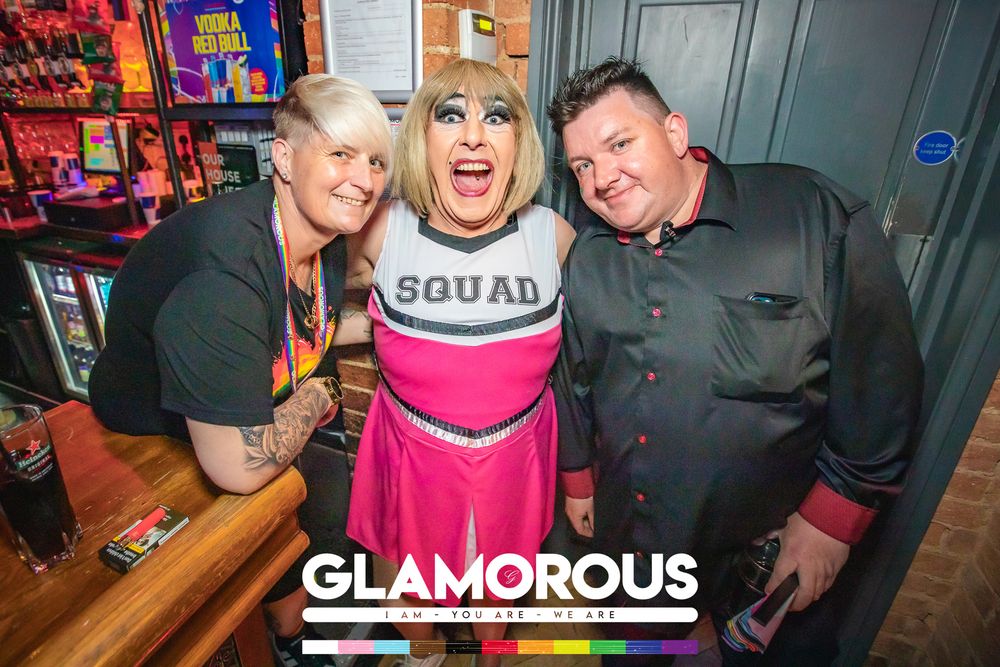 Three friends pose together at Glamorous nightclub, with center person in cheerleader costume reading Squad.
