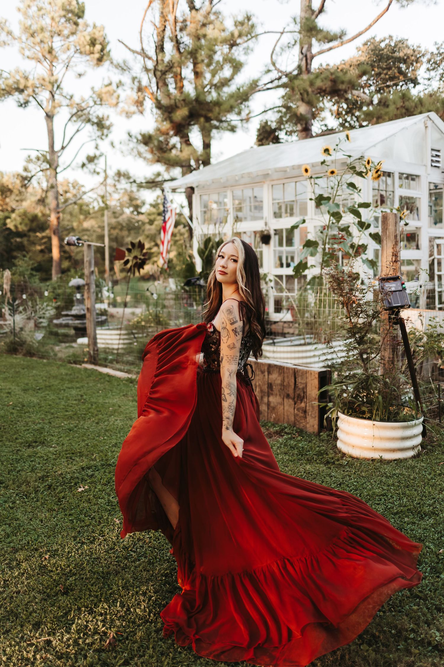 Woman in flowing red gown and cape poses gracefully in lush garden with greenhouse backdrop.