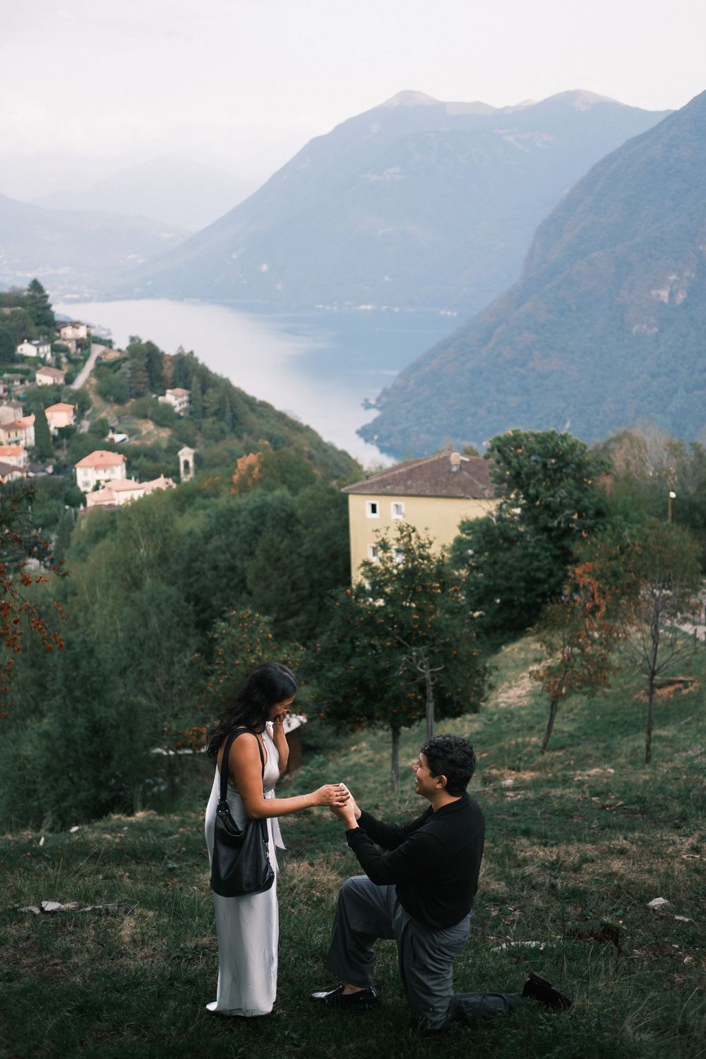 Man proposing on one knee to woman on a hillside with scenic lake and mountain views in background.