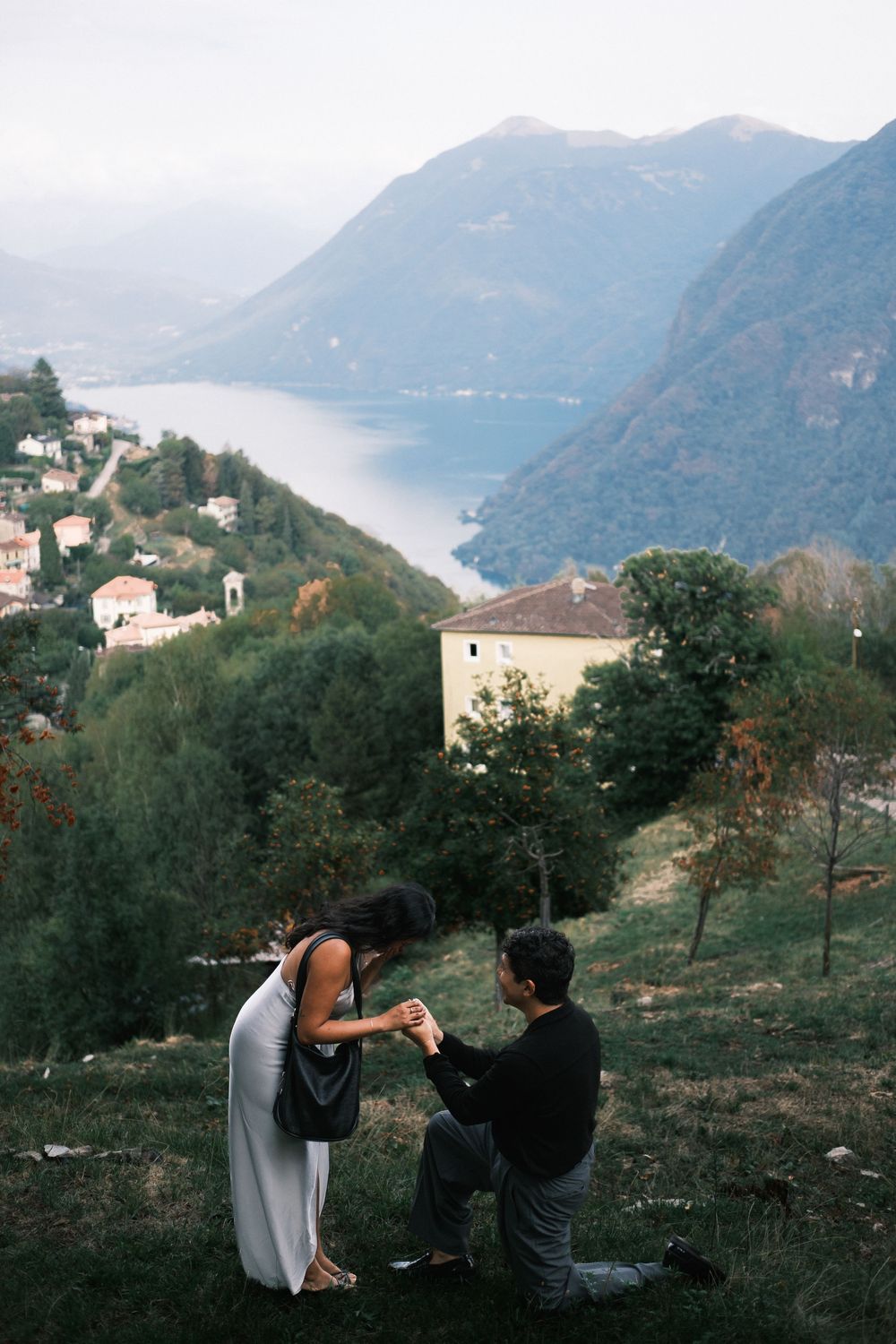 Man proposes to woman on hillside with stunning mountain lake backdrop in scenic European village setting.